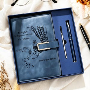 Op de afbeelding: Een blauw notitieboek met lederen kaft, voorzien van een Winnie de Poeh-illustratie en de tekst "Never forget the difference you make Mr. Johnson" en "Thank you James", in een geschenkdoos met een bijpassende blauwe pen en zilveren vulling.
