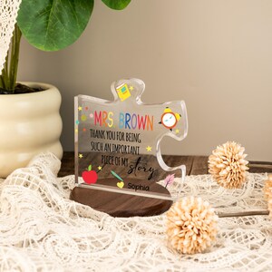 May include: A clear acrylic puzzle piece-shaped plaque with a wooden base. The plaque has colorful text that reads "MRS BROWN THANK YOU FOR BEING SUCH AN IMPORTANT PIECE OF MY STORY Sophia". The design includes a red apple, a pencil, and a clock.