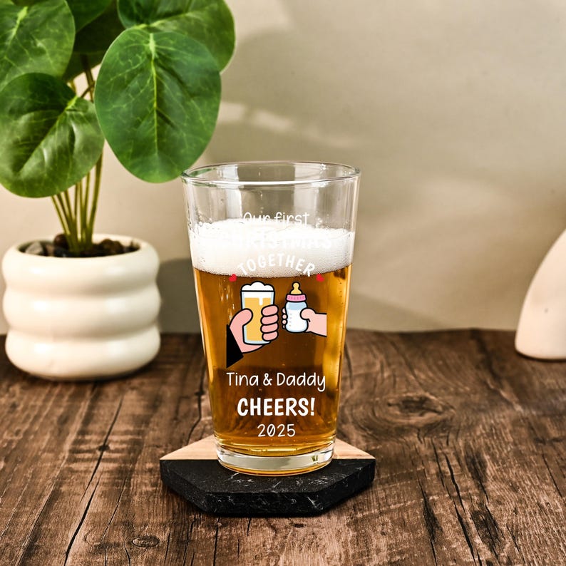 May include: A clear glass pint glass filled with golden beer and white foam. The glass features white text that reads Our first CHRISTMAS TOGETHER, Tina & Daddy CHEERS! 2025, and a graphic of a hand holding a beer glass clinking with a baby bottle.