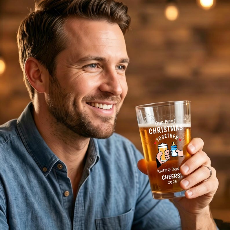 May include: A clear glass pint glass filled with amber liquid, featuring a printed graphic of a hand holding a beer and a hand holding a baby bottle. The text reads: Our first CHRISTMAS TOGETHER Keith & Dad CHEERS! 2025 with small red hearts.