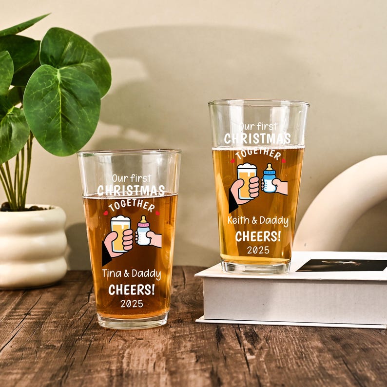 May include: Two clear glass pint glasses filled with amber liquid. Each glass features white text reading Our first CHRISTMAS TOGETHER, CHEERS! 2025, and a graphic of a hand holding a beer glass clinking with a hand holding a baby bottle.