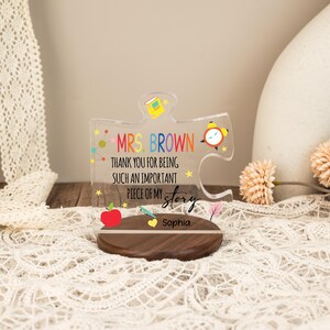 May include: A clear acrylic puzzle piece with the text "MRS. BROWN THANK YOU FOR BEING SUCH AN IMPORTANT PIECE OF MY story Sophia". The puzzle piece sits on a wooden base and features colorful illustrations.