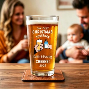 May include: A clear pint glass filled with amber beer, featuring white printed text that reads Our first CHRISTMAS TOGETHER, Keith & Daddy CHEERS! 2026, and a graphic of a hand holding a beer glass clinking with a hand holding a baby bottle.