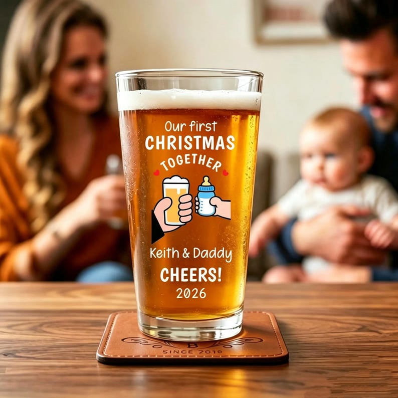 May include: A clear pint glass filled with amber beer, featuring white printed text that reads Our first CHRISTMAS TOGETHER, Keith & Daddy CHEERS! 2026, and a graphic of a hand holding a beer glass clinking with a hand holding a baby bottle.