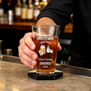 May include: A clear glass pint glass filled with an amber liquid, featuring the printed text Our first CHRISTMAS TOGETHER, Tina & Daddy CHEERS! 2026, and a graphic of a hand holding a beer glass clinking with a hand holding a baby bottle.
