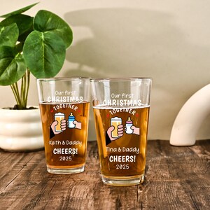 May include: Two clear glass pint glasses filled with amber liquid. Each glass features a graphic of a hand holding a beer and a hand holding a baby bottle, with the text Our first CHRISTMAS TOGETHER, names, CHEERS!, and the year 2025.