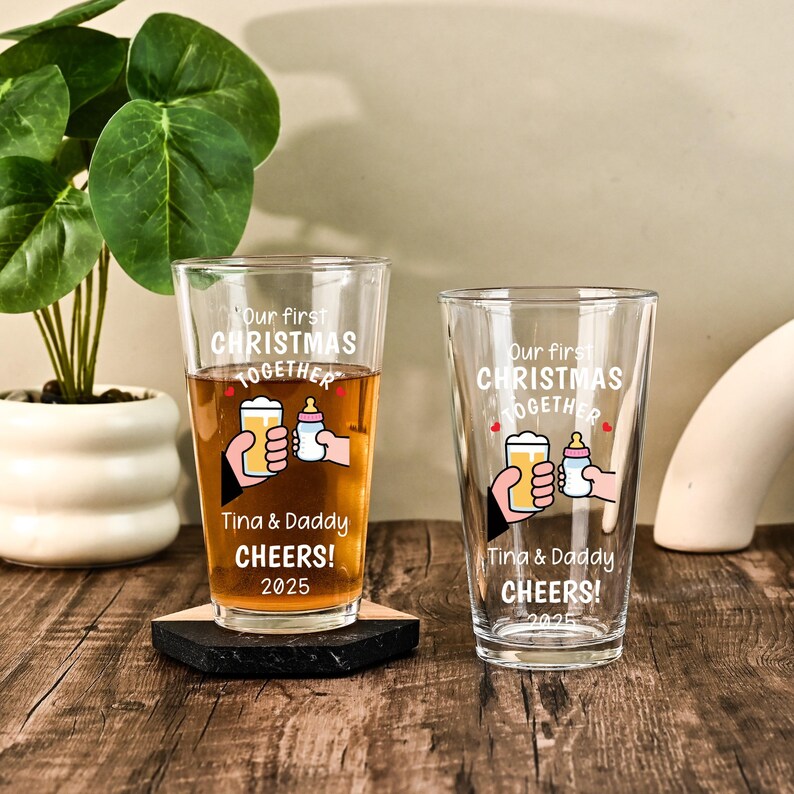 May include: Two clear glass pint glasses featuring the white printed text Our first CHRISTMAS TOGETHER, Tina & Daddy, CHEERS!, and 2025. The design includes a graphic of one hand holding a beer glass and another hand holding a baby bottle.