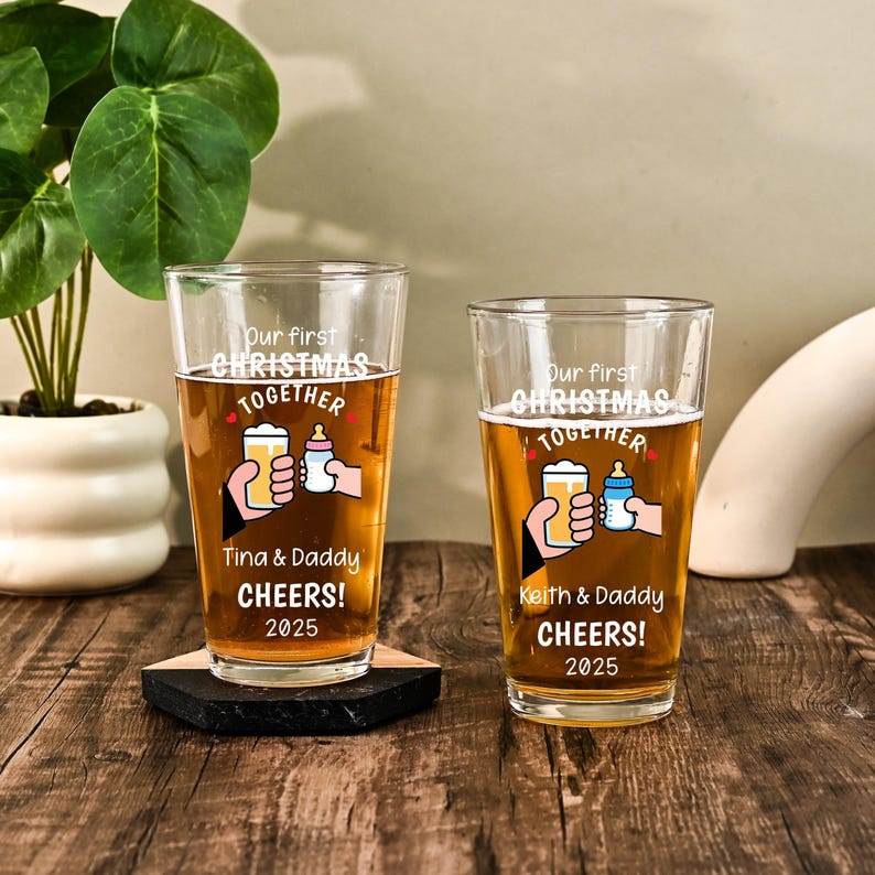 May include: Two clear glass pint glasses filled with amber liquid, each featuring white text that reads Our first CHRISTMAS TOGETHER, CHEERS! 2025, and a graphic of a hand holding a beer glass next to a hand holding a baby bottle with names.