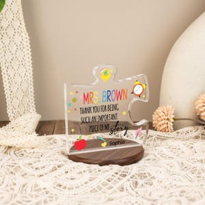 May include: A clear acrylic puzzle piece with the text "MRS. BROWN THANK YOU FOR BEING SUCH AN IMPORTANT PIECE OF MY story Sophia". The puzzle piece has colorful illustrations of a book, an alarm clock, and an apple. It sits on a wooden base.