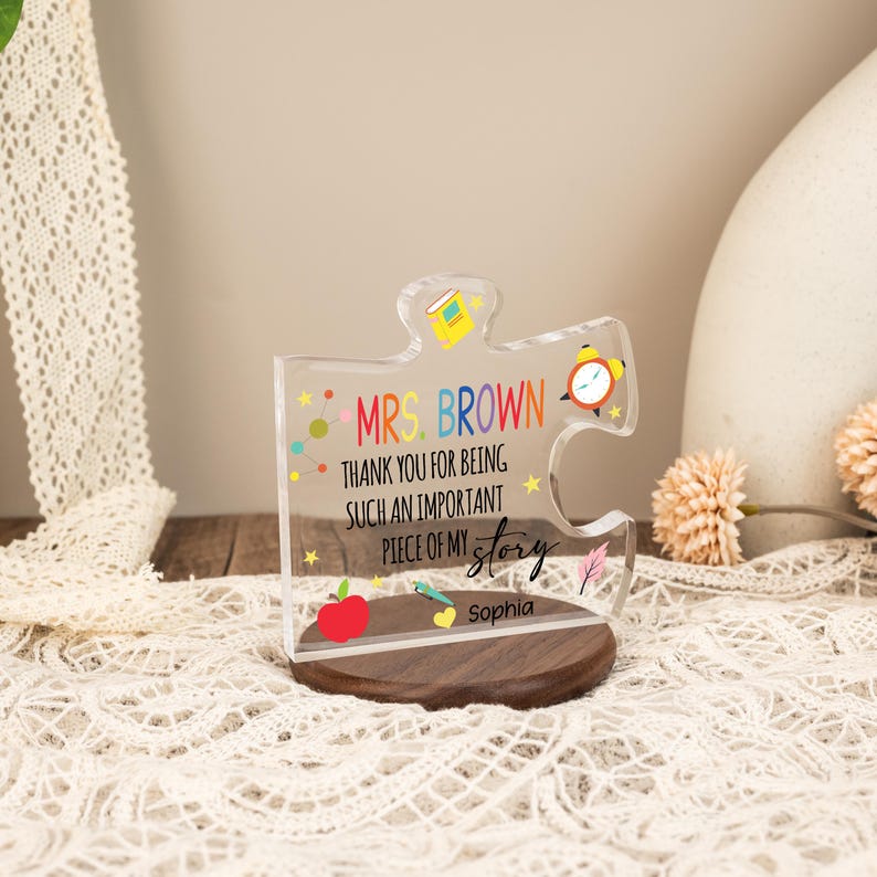 May include: A clear acrylic puzzle piece with the text "MRS. BROWN THANK YOU FOR BEING SUCH AN IMPORTANT PIECE OF MY story Sophia". The puzzle piece has colorful illustrations of a book, an alarm clock, and an apple. It sits on a wooden base.