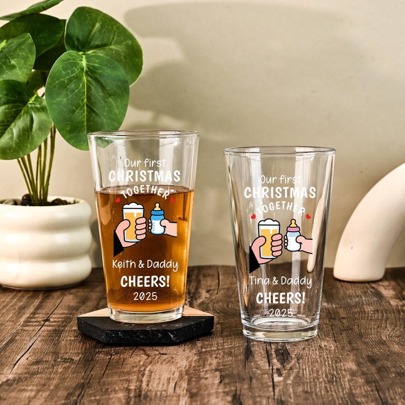 May include: Two clear glass pint glasses featuring white text that reads Our first CHRISTMAS TOGETHER, CHEERS! 2025, and names Keith & Daddy or Tina & Daddy. The design includes a graphic of a hand holding a beer glass and a baby bottle.