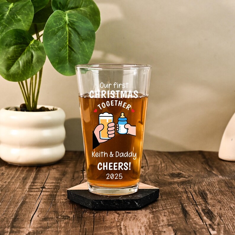 May include: A clear glass pint glass filled with amber liquid, featuring white text that reads Our first CHRISTMAS TOGETHER, Keith & Daddy CHEERS! 2025, and a graphic of two hands clinking a beer glass and a baby bottle together.