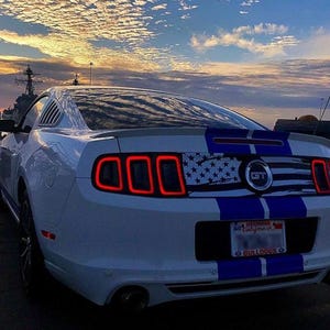 May include: White Ford Mustang GT with a blue and white American flag decal on the back. The car has a blue stripe down the side and a "Georgia to California" license plate with "Bulldogs" on the bottom.