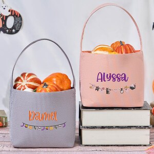 Puede incluir: Dos bolsas de mano con tem&aacute;tica de Halloween, una a rayas blancas y negras, la otra naranja y blanca. Cada bolsa est&aacute; llena de calabazas y tiene un nombre impreso en la parte delantera. Una pancarta con decoraciones de Halloween est&aacute; debajo de los nombres.