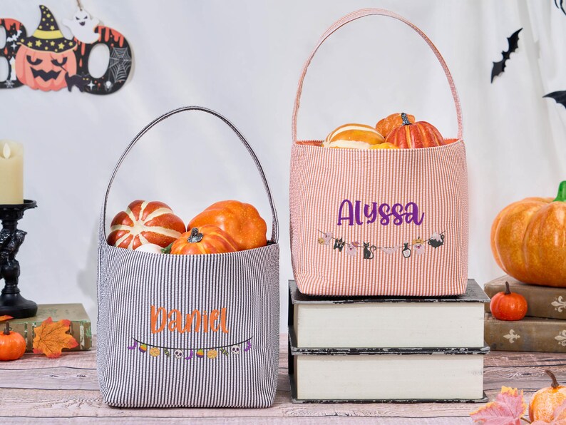Puede incluir: Dos bolsas de mano con tem&aacute;tica de Halloween, una a rayas blancas y negras, la otra naranja y blanca. Cada bolsa est&aacute; llena de calabazas y tiene un nombre impreso en la parte delantera. Una pancarta con decoraciones de Halloween est&aacute; debajo de los nombres.
