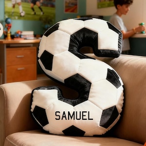 Puede incluir: Un gran cojín con forma de letra "S" diseñado para parecerse a un balón de fútbol, con paneles pentagonales en blanco y negro. El nombre "SAMUEL" está impreso en negro en la sección inferior. El cojín está sobre un sofá beige.