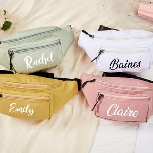 May include: Four personalized fanny packs in pastel colors: sage green, white, mustard yellow, and pink. Each pack has a front pocket and a zipper closure. Names "Rachel", "Baines", "Emily", and "Claire" are printed on the front.