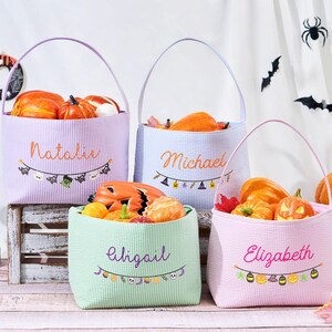 Puede incluir: Cuatro bolsas de golosinas de Halloween en colores pastel llenas de calabazas. Cada bolsa tiene un asa y un nombre bordado en la parte delantera: Natalie, Michael, Abigail y Elizabeth. Una pancarta festiva con iconos de Halloween decora cada bolsa.