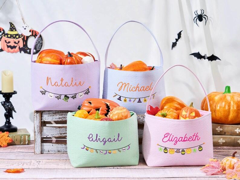 Puede incluir: Cuatro bolsas de golosinas de Halloween en colores pastel llenas de calabazas. Cada bolsa tiene un asa y un nombre bordado en la parte delantera: Natalie, Michael, Abigail y Elizabeth. Una pancarta festiva con iconos de Halloween decora cada bolsa.