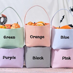 Puede incluir: Seis coloridas bolsas de golosinas de Halloween de tela seersucker en verde, naranja, azul, morado, negro y rosa. Cada bolsa est&aacute; llena de calabazas decorativas. Las bolsas tienen un asa y el nombre del color est&aacute; impreso.