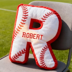May include: A baseball-style cushion shaped like the letter 'R', with the name 'ROBERT' printed on it. The cushion is white with red stitching and a red border, mimicking a baseball. It is placed on a grey chair.