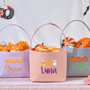 Puede incluir: Tres bolsas de mano con tem&aacute;tica de Halloween con asas, llenas de calabazas y objetos decorativos. Las bolsas son a rayas en morado, naranja y gris, cada una con un nombre y gr&aacute;ficos de Halloween. Las bolsas est&aacute;n sobre una superficie de madera.