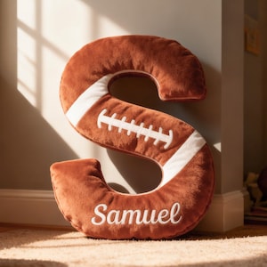 May include: A plush, brown football-shaped letter "S" pillow with white stripes and stitching. The name "Samuel" is embroidered in white script. The pillow is on a light-colored carpet.