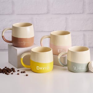 以下が含まれることがあります： ツートンカラーのデザインのセラミックマグカップ4つ。各マグカップはクリーム色のトップと、茶色、ピンク、黄色、緑の色合いのカラーボトムを備えています。各マグカップには名前が書かれています：アリッサ、エラ、デビッド、ウィリアム。