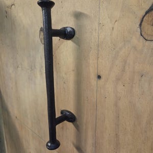 May include: A black, wrought iron door pull handle. The handle is mounted on a light brown wooden surface. The handle has a vertical bar with two horizontal bars extending from it. The handle is a dark, matte black color.