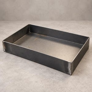May include: A rectangular metal tray with a dark grey finish and visible welded seams at the corners. The tray features high, straight sides and a flat base, showcasing a raw, industrial appearance with subtle variations in the metal surface texture.