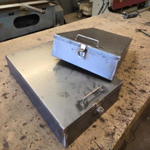 Könnte beinhalten: Zwei rechteckige Metallaufbewahrungsboxen mit roher Stahloberfläche. Jede Box verfügt über einen Klappdeckel, einen oben angebrachten Metallgriff und einen vorderseitigen Verschlussmechanismus. Die Boxen sind gestapelt, mit einer größeren Basis und einem kleineren Deckel.