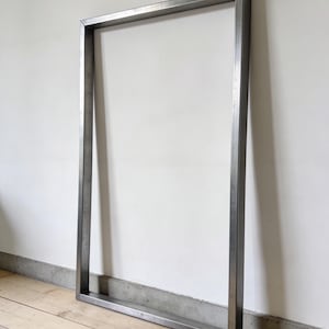 Puede incluir: Una estructura rectangular de marco abierto hecha de metal de color plateado. El marco está apoyado contra una pared blanca y descansa sobre un suelo de madera. El marco de metal es de diseño industrial simple.