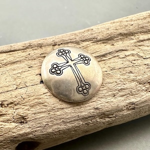 May include: A circular silver-toned metal button featuring a stamped cross design with trefoil ends. The cross is centered on the smooth, slightly domed surface of the button, which has a metallic finish and dark recessed lines defining the cross.