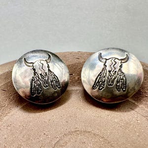 May include: A pair of circular silver-toned metal buttons or conchos featuring a stamped design of a bull skull with horns and hanging feathers. The metal surface has a polished finish with darkened recessed areas highlighting the engraved details.