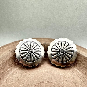 May include: A pair of silver-toned stud earrings. Each earring features a central circular design with radiating lines, surrounded by a scalloped edge. The earrings are displayed on a brown, textured surface.