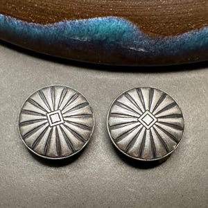 May include: A pair of silver-toned stud earrings. Each earring is round with a central square and radiating lines. The earrings are set against a neutral background.