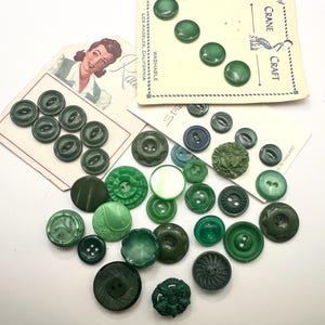 May include: A collection of various green buttons in different shades, sizes, and textures, including several attached to vintage cardboard display cards with printed text like Crane Craft and Los Angeles, California, scattered on a white surface.