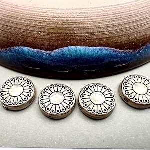 May include: Four small, round, silver-colored containers with a floral design. The containers are arranged in a row in front of a large, decorative bowl with brown, blue, and white colors.
