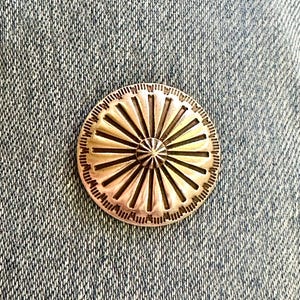 May include: A silver-colored, round metal button with a sunburst design. The button features a central, star-shaped detail and radiating lines. The button is set against a denim background.