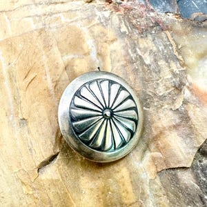 May include: A circular silver button featuring a raised, sunburst-like pattern with radiating petals extending from a central point. The metal surface has a polished finish with darkened recessed areas between the petals to highlight the design.