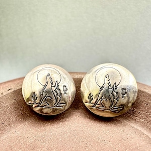 May include: Pair of round stud earrings with a silver wolf howling at the moon design. The earrings have a desert theme with cacti and a full moon. The earrings are set on a light-colored background.