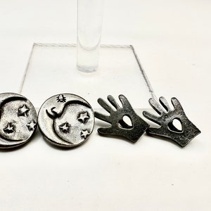 May include: Three silver-toned metal earrings. Two earrings feature a crescent moon design with stars. One earring features a hand with a heart cutout.