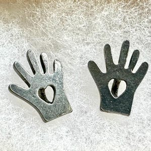 May include: A pair of silver-toned stud earrings shaped like stylized hands with five fingers. Each earring features a small, heart-shaped cutout in the center of the palm area, providing a distinct and symmetrical design for these metallic accessories.