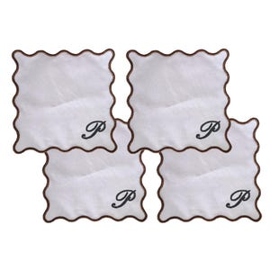 Könnte beinhalten: Vier weiße quadratische Untersetzer mit einem gewellten braunen Rand. Jeder Untersetzer hat einen schwarz gestickten Schriftzug "P" in der Mitte. Die Untersetzer sind leicht überlappend angeordnet.