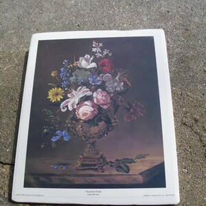 May include: A vintage print of a still life featuring a vase filled with a variety of flowers, including white lilies, pink roses, and blue forget-me-nots. The vase is ornate and gold in color. The background is a dark brown.