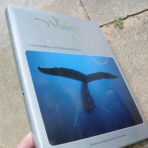May include: A book titled "Whales of Hawaii" with a picture of a whale's tail underwater. The cover is silver with green text and the title. The book is about the pictorial history of whaling and Hawaii.