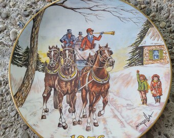 May include: A round decorative porcelain plate with a gold rim featuring a winter scene of a horse-drawn carriage with three passengers, one blowing a horn. Two children stand in the snow nearby. The year 1976 is printed at the bottom.