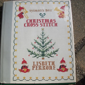 May include: A vintage "Woman's Day Christmas Cross Stitch" book. The cover features a cross-stitched Christmas tree, festive figures, and text. The book is bound with a white spine and a green base.