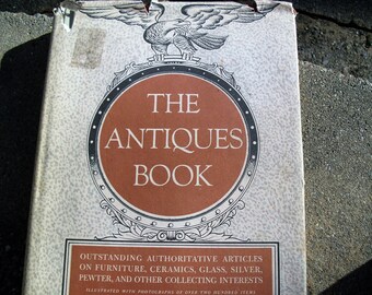 May include: The Antiques Book, a vintage hardcover book with a decorative cover. The cover features a brown circular design with the title "THE ANTIQUES BOOK" and an eagle illustration. The book's cover also includes text about furniture, ceramics, glass, silver, pewter, and other collecting interests.
