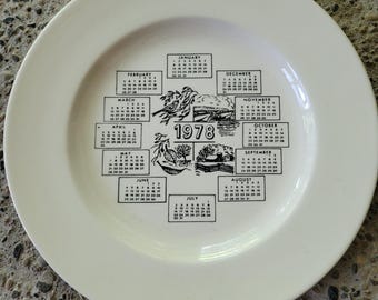 May include: A round, off-white ceramic plate featuring a black printed calendar for the year 1978. The design includes twelve monthly grids arranged in a circle around the central year 1978, accompanied by small illustrations of birds and nature.